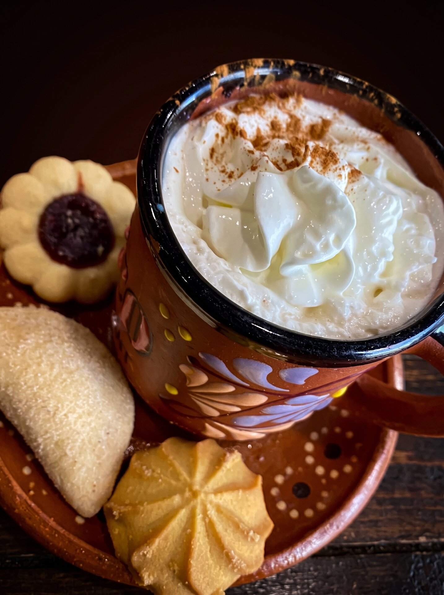 Champurrado topped with cream in a painted clay mug, beside three pieces of pan dulce